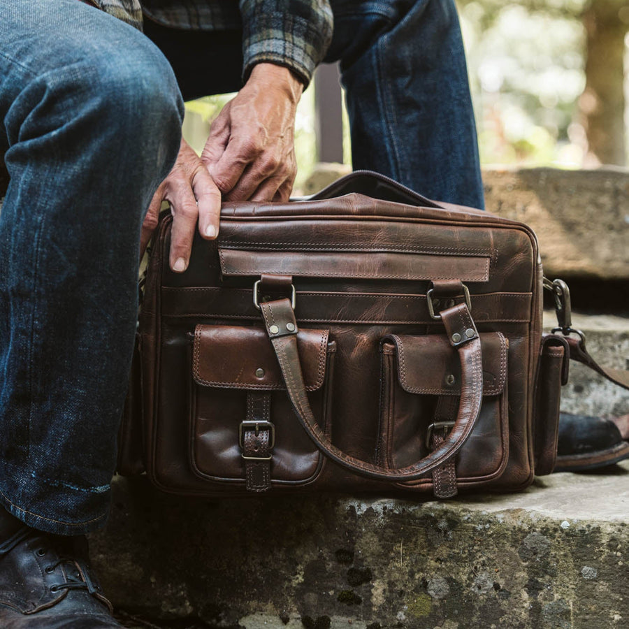 Men's rugged leather pilot bag in dark oak with multiple compartments, brass buckles, adjustable strap, and detachable pouches. Ideal for travel.